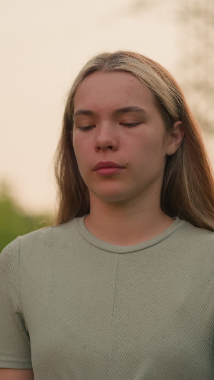 primer plano de una mujer joven con expresión cansada y contemplativa caminando al aire libre, vestida con una camisa verde casual, en un fondo natural y borroso de vegetación en una luz suave