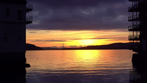 Golden-sunset-above-Askoy-island-outside-Bergen-seen-in-between-silhouette-of-two-apartment-buildings-in-Sandviken---Beautiful-sun-reflections-in-calm-sea