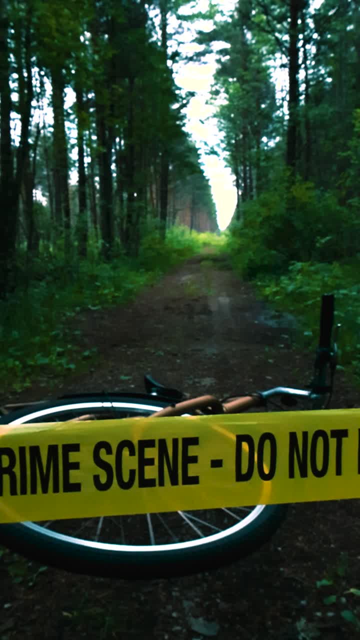 Scary footage of a vibrant yellow Crime Scene Do Not Enter tape ribbon pulled in front of a bicycle that is laying on a muddy forest road ground during a cloudy day, person is missing, investigation.