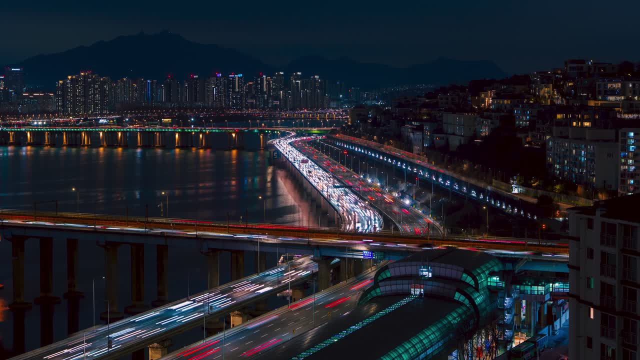 Timelapse che mostra continue scie luminose del traffico notturno che scorrono lungo la superstrada Gangbyeonbuk-ro e il Ponte Donho sul fiume Han, circondato da grattacieli dello skyline di Seul e riflessi.