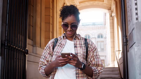 Primer-Plano-De-Una-Joven-Negra-De-Moda-Con-Gafas-De-Sol-Y-Camisa-A-Cuadros-Usando-Su-Teléfono-Inteligente-Parado-Afuera-De-La-Entrada-De-Un-Edificio-Histórico,-En-ángulo-Bajo