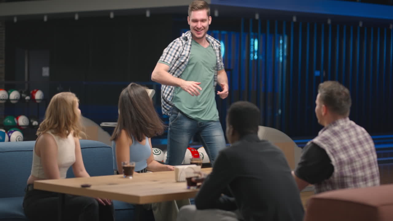 Un uomo gioioso corre verso gli amici dando il cinque a uomini e donne seduti a un tavolo in una sala da bowling. Celebrano la vittoria e la serie positiva. Per congratularsi della vittoria. Successo di squadra