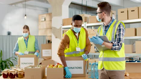 Front-view-of-caucasian-male-volunteer-in-facial-mask-checking-donation-list-and-talking-with-his-coworkers