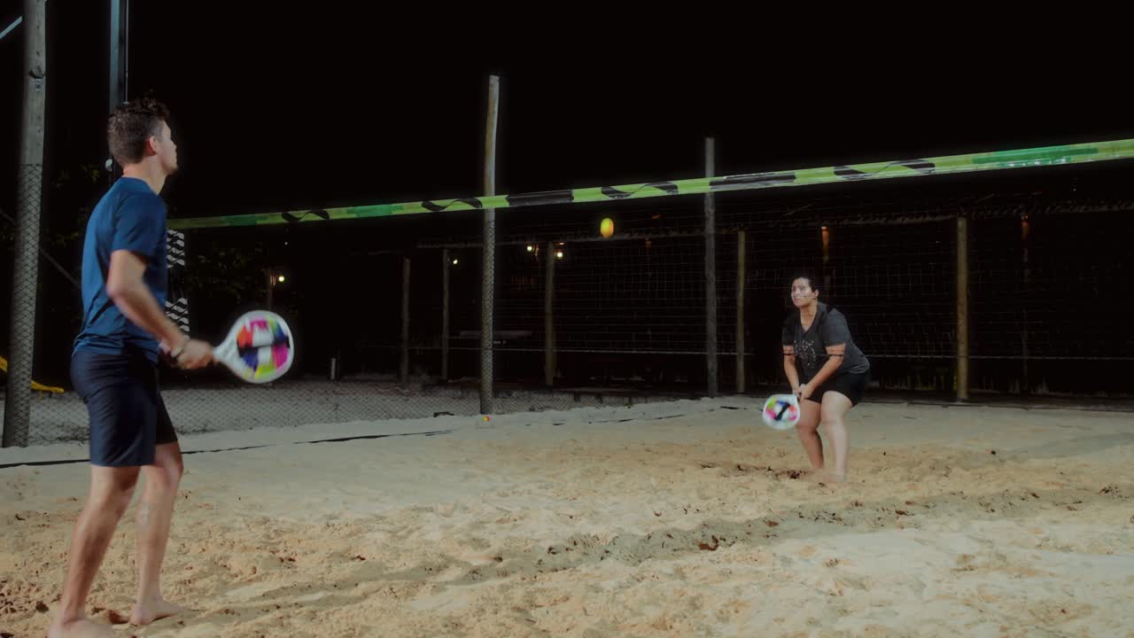 Two people playing beach tennis or padel on a sand court at night