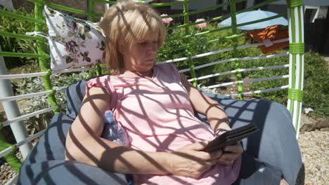 blonde woman in shorts reads ebook novel relaxing in comfy garden hammock on hot sunny day