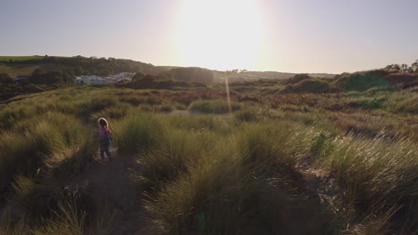 Drohnenaufnahme-Eines-Jungen-Mädchens-Im-Strandurlaub,-Das-In-Sanddünen-Vor-Der-Flackernden-Sonne-Spielt