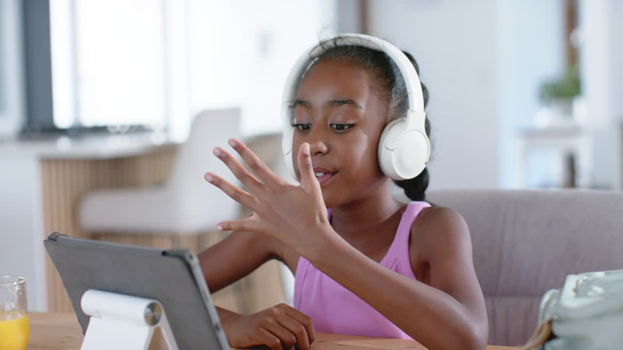 Afrikanisch-amerikanisches Mädchen zu Hause mit Headset, das auf eine Lehreraufforderung reagiert, sich vorbeugt und auf ein Tablet tippt.