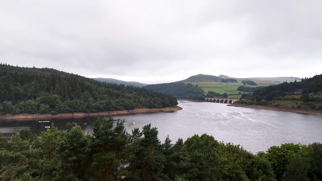 Aerial drone footage glides above a wide reservoir bordered by dense forest and rolling hills under overcast skies, revealing a distant dam structure