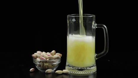 Beer-is-pouring-into-glass-on-black-background.-Bowl-of-pistachios-nuts