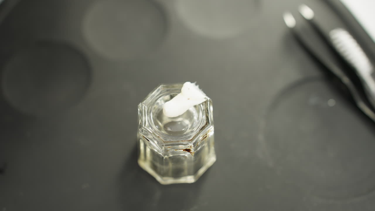 Close-up view of technician wearing black gloves squeezing thick white cream from tube into small clear glass container placed on black silicone board with beauty tools scattered around during setup