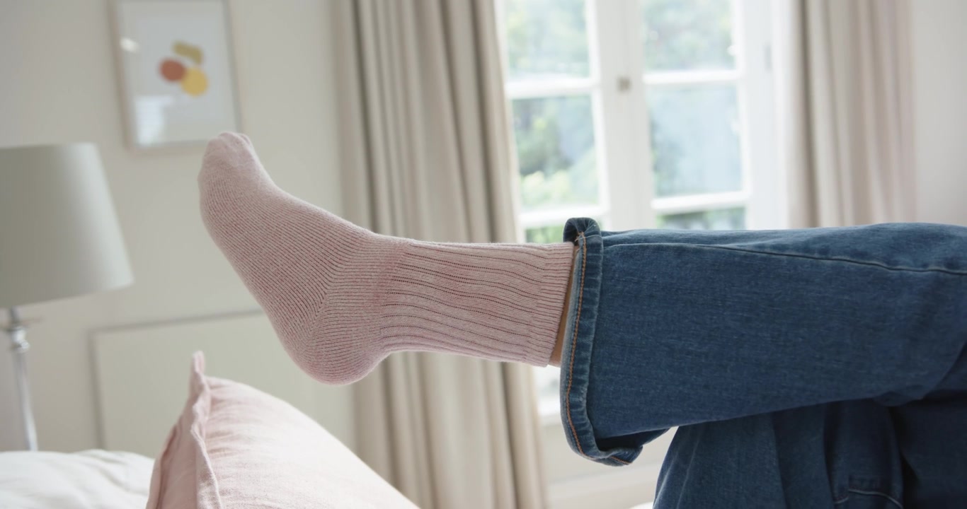Una donna afroamericana sdraiata sul letto solleva e flette un piede con il calzino, indossando jeans risvoltati, in cerca di riposo.