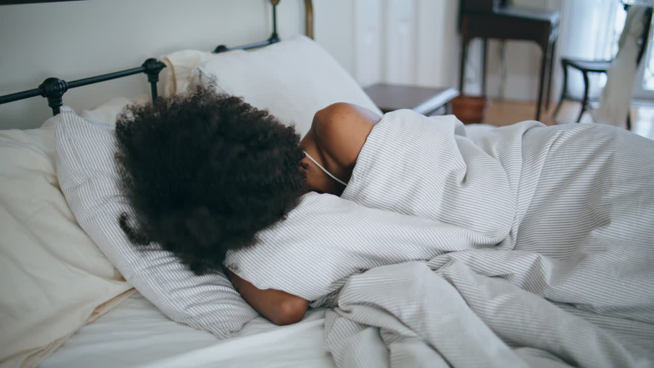Træt pige ligger i sengen om morgenen. Krølhåret, doven kvinde vågner op i et hyggeligt værelse.
