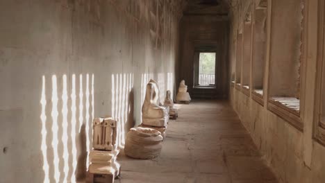 forward dolly inside a hallway with headless statues and light shining from the windows, inside angkor wat.
