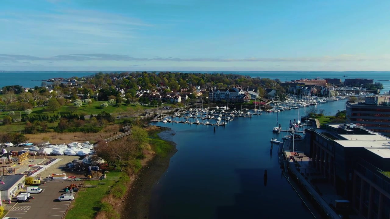 Ripresa con drone che inquadra dal lungomare verso un porto turistico, un'area residenziale e l'orizzonte costiero durante una giornata primaverile a Stamford, Connecticut