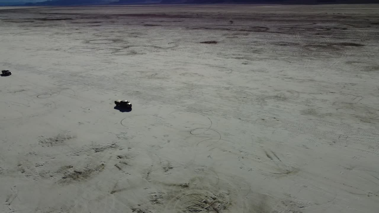 Two trucks driving in a caravan on the Bonneville Salt Flats in Utah. Aerial shot revealing tire marks and treads from other vehicles. White salt contrasted by the trucks. 4K cinematic drone shot.