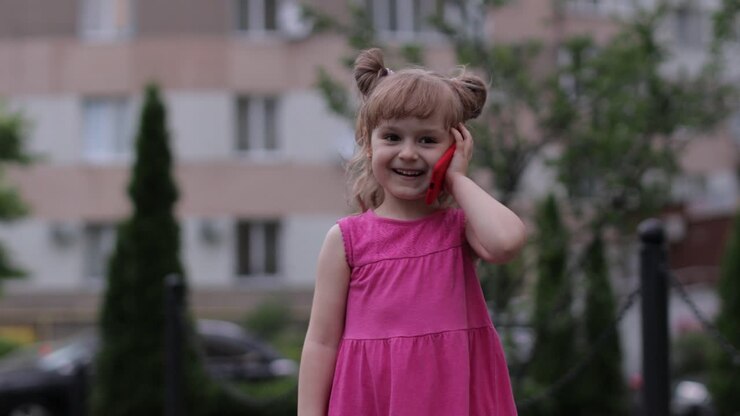 Child girl talking by smartphone outdoors. Kid in pink dress talking by mobile phone on city street