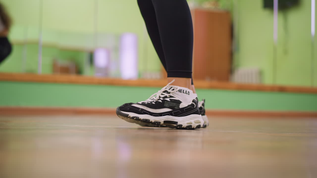 Leg view of girl in black leggings and canvas sneakers bouncing on polished wooden floor during workout session, capturing rhythm, energy, and athletic movement in blurred background of indoor studio