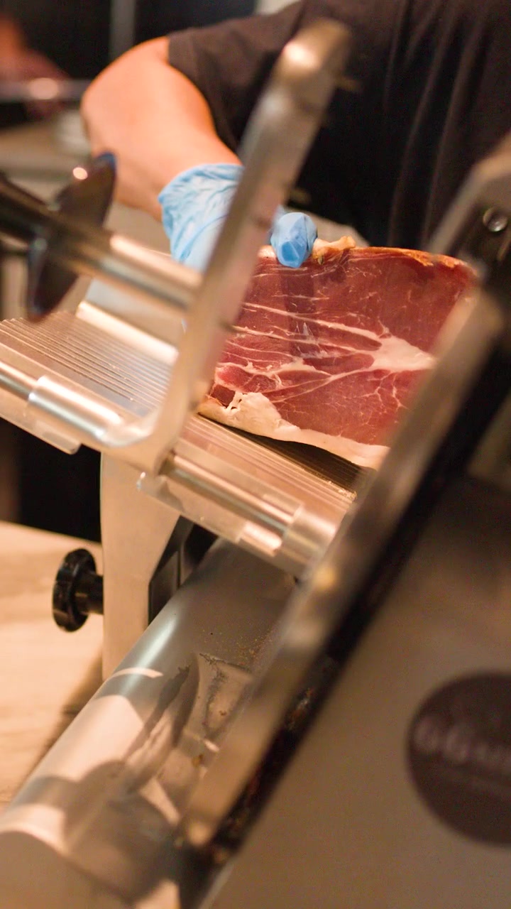 Close-up of chef using electric slicer on cured meat in a professional restaurant kitchen.