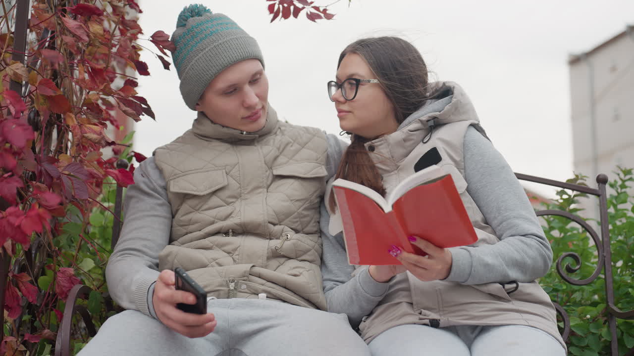 Romantic moment of lovers seated on park bench in autumn, woman reading book while man hugs her warmly holding phone, both dressed in cozy jackets, surrounded by red leaves and greenery