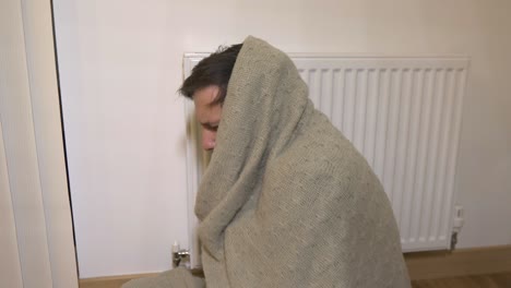 freezing young man wrapped in blanket sitting next to a heater and shivering from cold, energy crisis