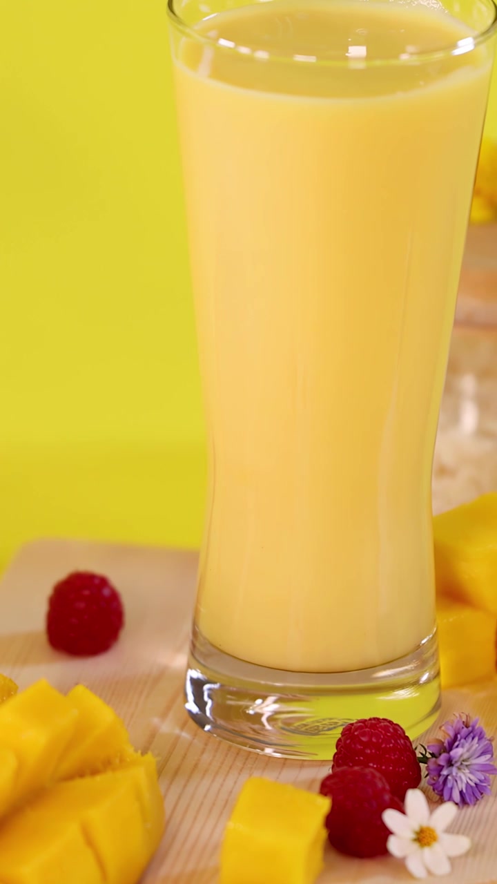 Smoothie al mango versato in un bicchiere accanto a mango a cubetti, lamponi e fiori su sfondo giallo brillante.