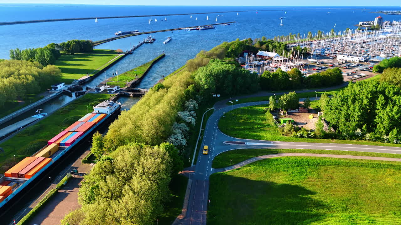 Large barge loaded with containers stands over the bridge at the dam. Flight over the green waterfront of Lelystad, the Netherlands.