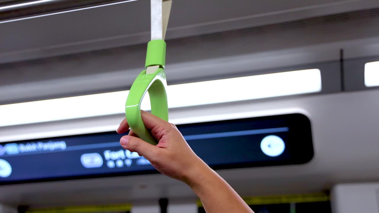 A person’s hand firmly holds a green strap handle inside a brightly lit, modern subway train, with a digital station sign visible in the background
