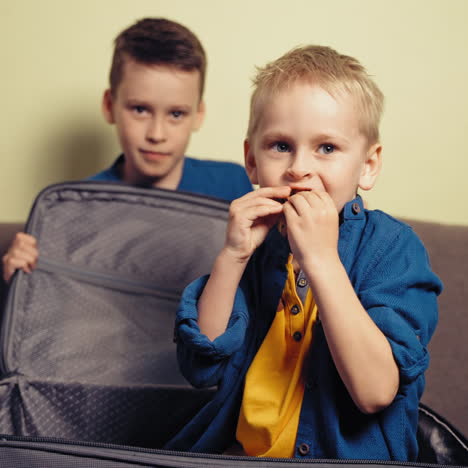 cheerful brothers playing at home with grey valise on the sofa. a boy opens the big suitcase and a little boy gets out inside of it and eat something.