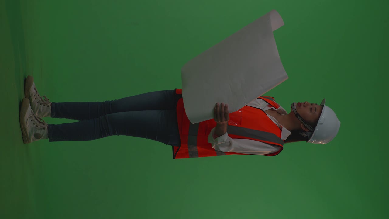 Side View Of Full Body Asian Female Engineer With Safety Helmet Looking At Blueprint In Her Hands And Looking Around While Standing In The Green Screen Background Studio, During Sunset Or Sunrise Time