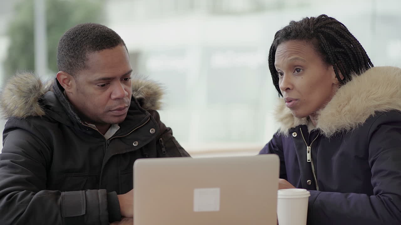 Afro-American middle-aged man in black jacket with fur hood discussing new project idea with Afro-American serious young girl with braids outside