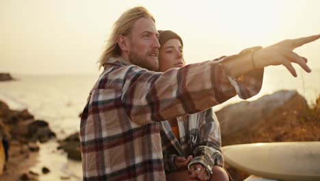 a blond man with a beard in a plaid shirt sits next to his blond girlfriend in a hat and a plaid shirt, he points somewhere to the side and they laugh on the rocky earthen shore of the sea at sunrise in the summer, their surfboards lie near them