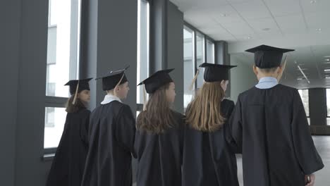 back view of kindergarten students in cap and gown holding graduation diploma and walking in the school corridor