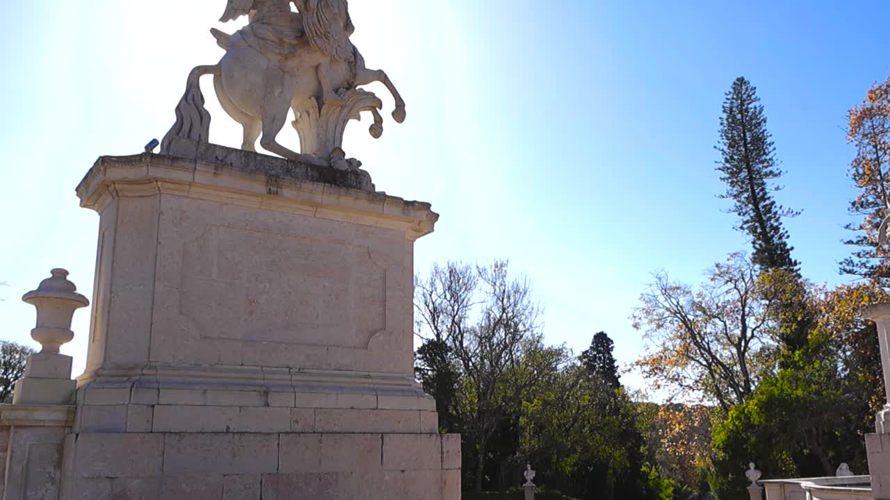Une statue équestre rétroéclairée se dresse sur un ciel bleu clair tandis que la lumière du soleil découpe la pierre sculptée et la verdure environnante, créant ainsi une scène de jardin spectaculaire et élégante.