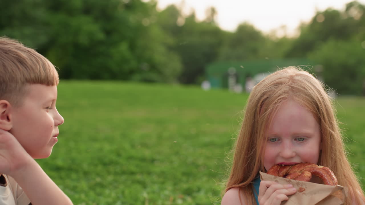 two children sitting on picnic mat in park green grass summer day eating fries, smiling and chatting, relaxed outdoor snack time with soft background trees and warm light