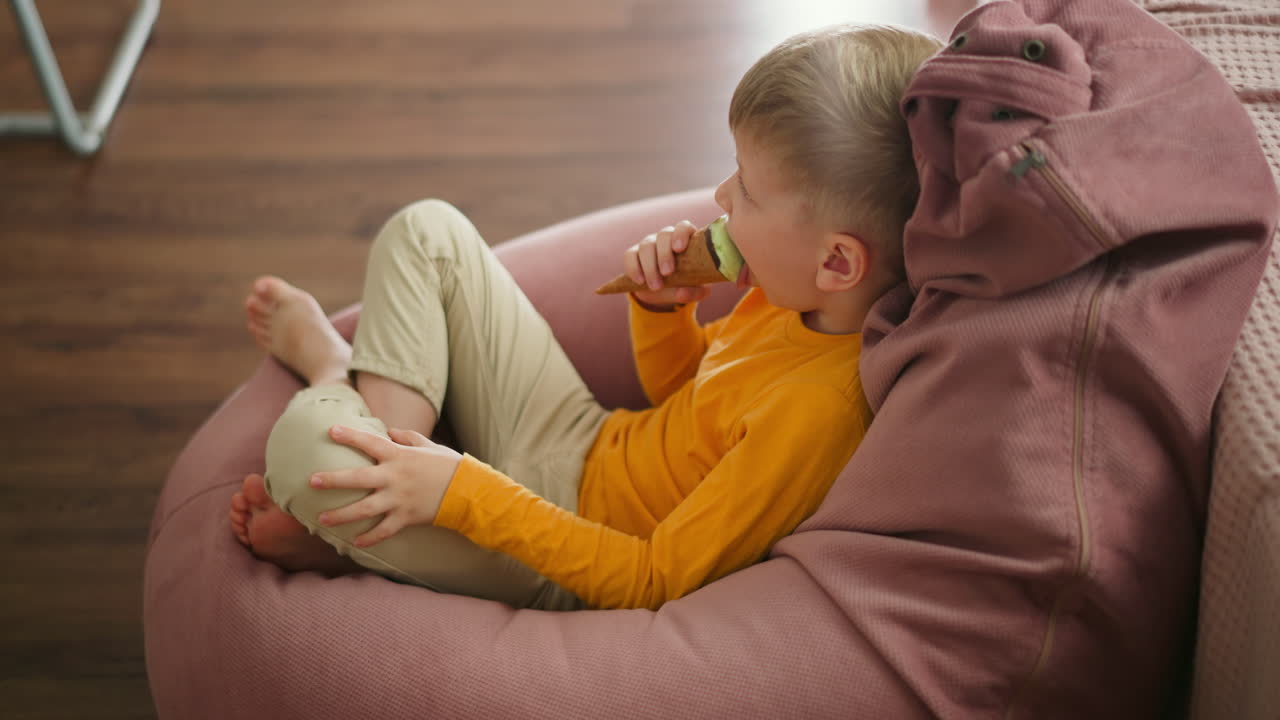 Young boy sitting comfortably on large pink beanbag licking mint ice cream cone while looking thoughtfully at background where part of metal stand is visible