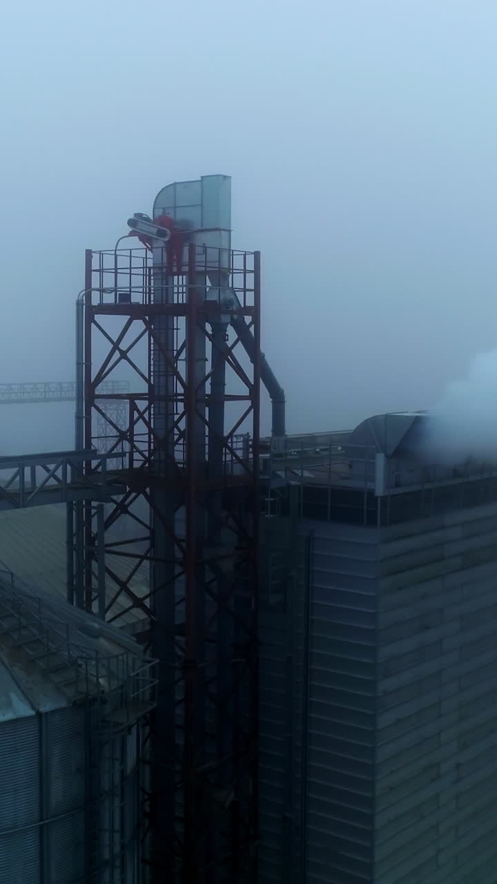 Approaching the high building at elevator plant emitting thick white smoke. Aerial view. Dirty tops of granary tanks at agribusiness enterprise. Foggy day backdrop. Vertical video