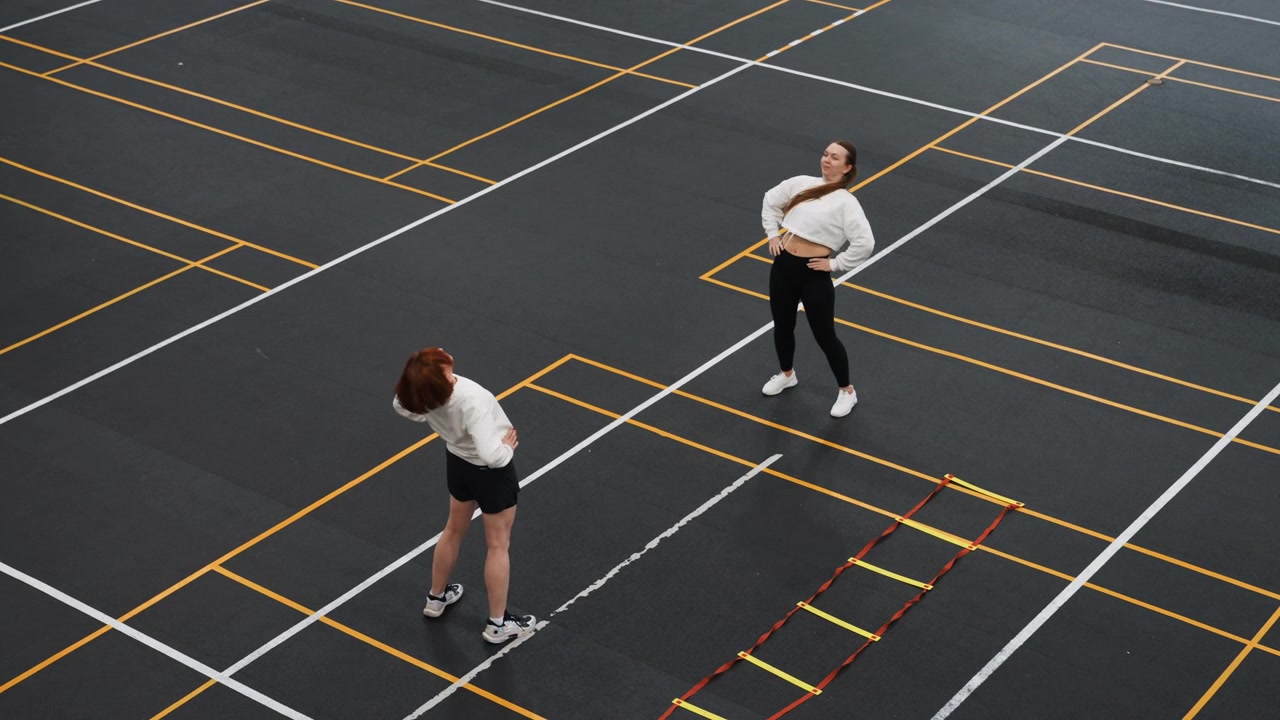 Two players practicing badminton on asphalt, coach guiding footwork beside agility ladder, aerial view of marked training grid showing racket swings, focused warmup and precision step drills