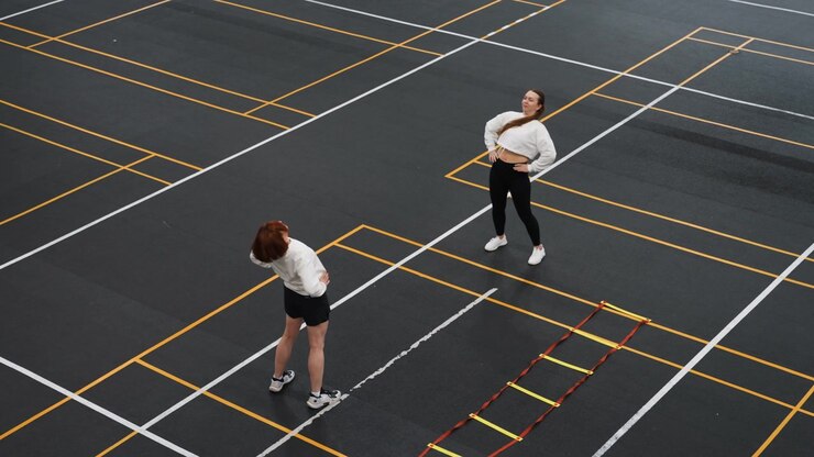 Two players practicing badminton on asphalt, coach guiding footwork beside agility ladder, aerial view of marked training grid showing racket swings, focused warmup and precision step drills
