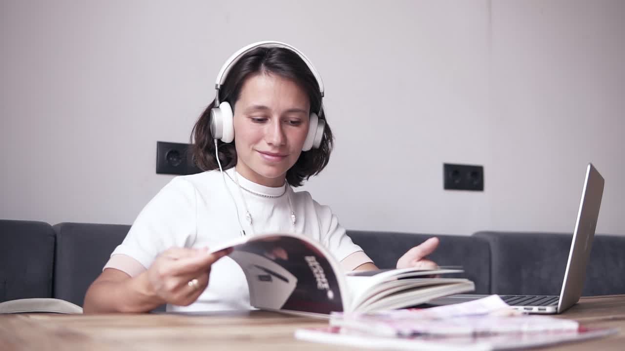 chica bonita disfruta de su tiempo mientras mira a través, leyendo la revista. se sienta junto a la mesa y escucha la música en auriculares. relajado. en el interior