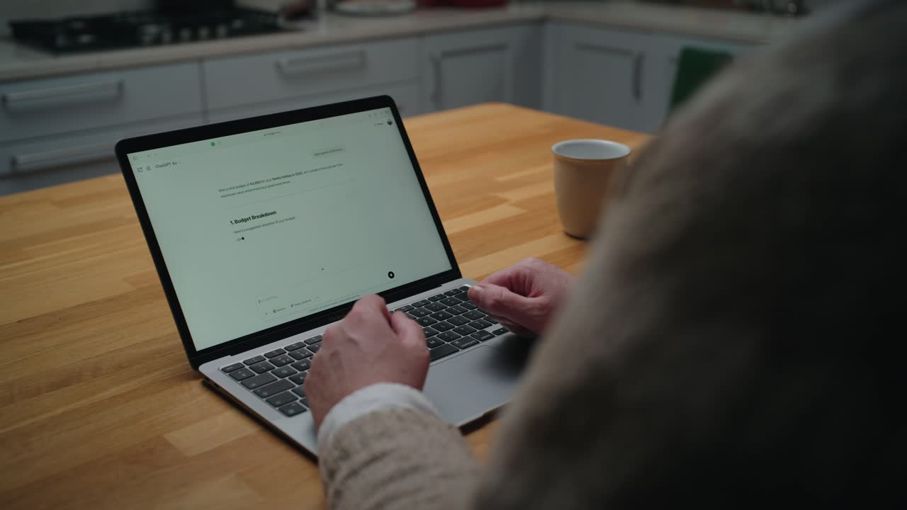 A close-up of hands typing on a laptop displaying an AI chatbot calculating a travel budget. Cozy kitchen setting with a wooden table and coffee cup in the background.