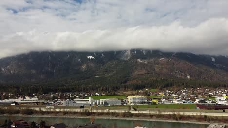 Drone-flies-up-over-Brixlegg-in-Tyrol,-mountains-with-clouds-and-the-river-Inn