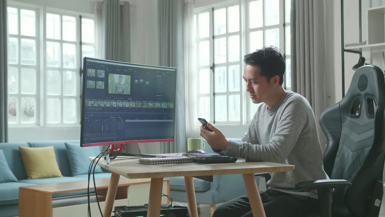 Asian Video Editor Man In Long Sleeved T-Shirt And Black Pants Using Mobile Phone While Using Desktop Computer For Editing Video At Home.