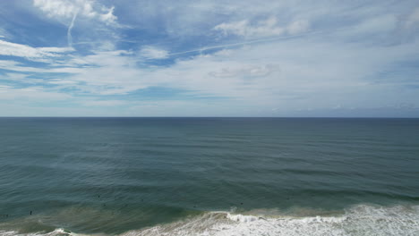 surfers catch waves under a vast blue sky along a serene, expansive coastline