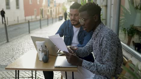 multiethnic men working with papers and laptop in cafe