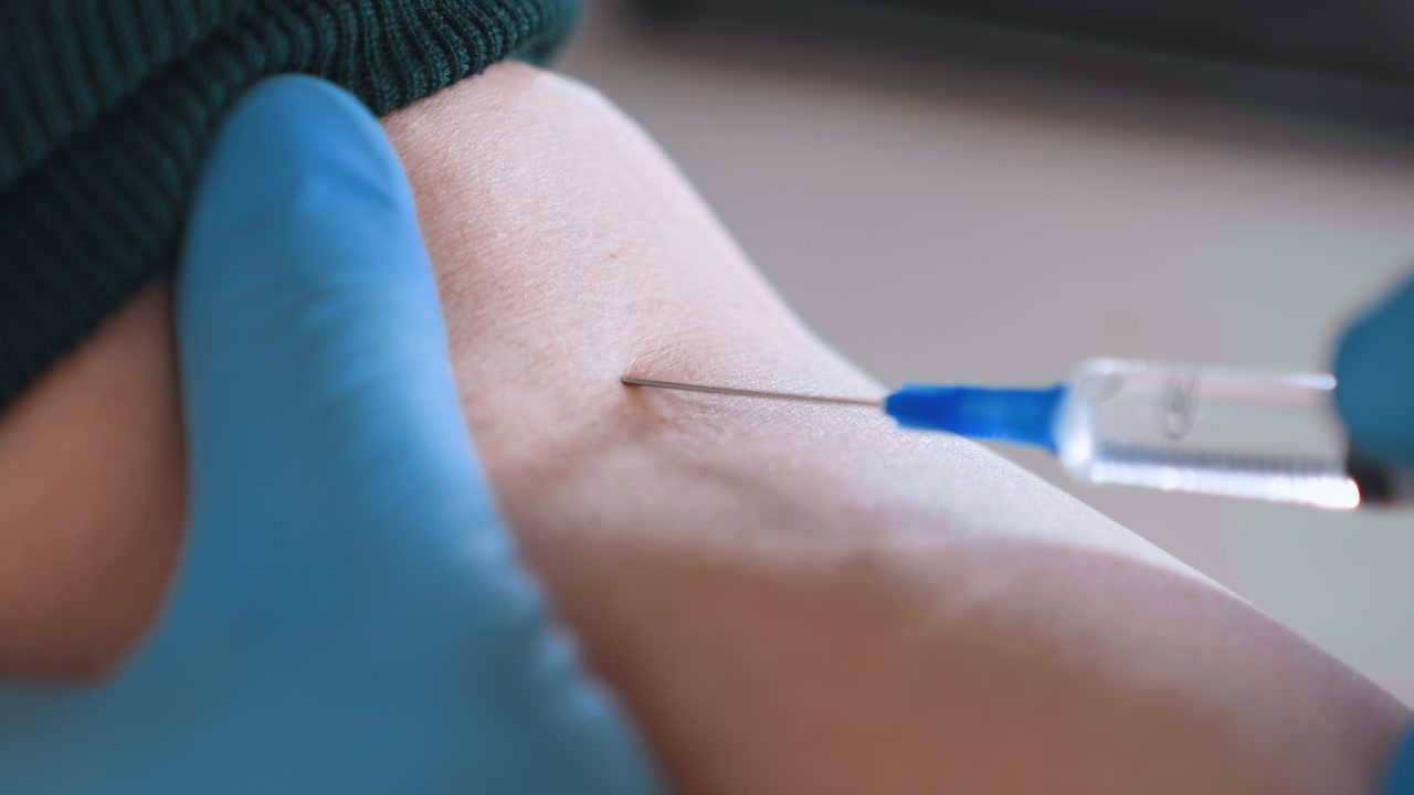 Nærbillede af arm, der modtager injektion af sygeplejerske, blå handske stabiliserer indre albue, sprøjte gennemborer hud til vaccination, antiseptisk forberedelse, hjemligt interiør, blid, rolig hånd, professionel pleje