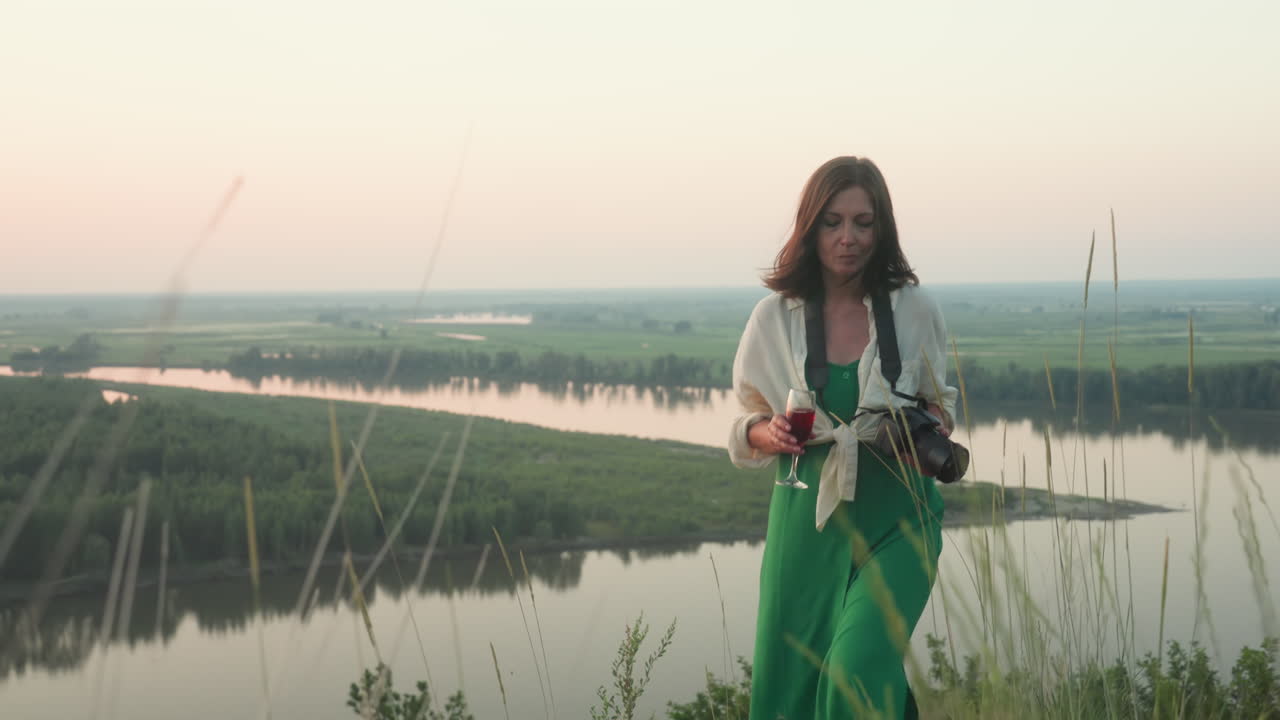 woman climbing grassy hill holding wine glass and camera under soft sunset light scenic background features calm river winding through lush forest and open field horizon
