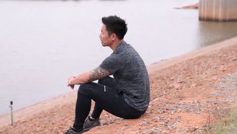 asian male sits on hill near the lake looking out to the water