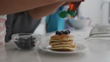 Midsection-of-girl-in-kitchen,-putting-syrup-on-stack-of-pancakes