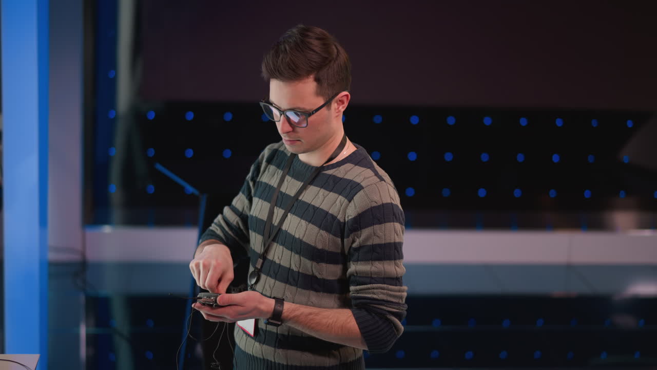 Male technician wearing glasses and striped sweater monitors wireless microphone transmitter and connects cables using handheld audio device inside modern television studio illuminated by blue white backdrop lights