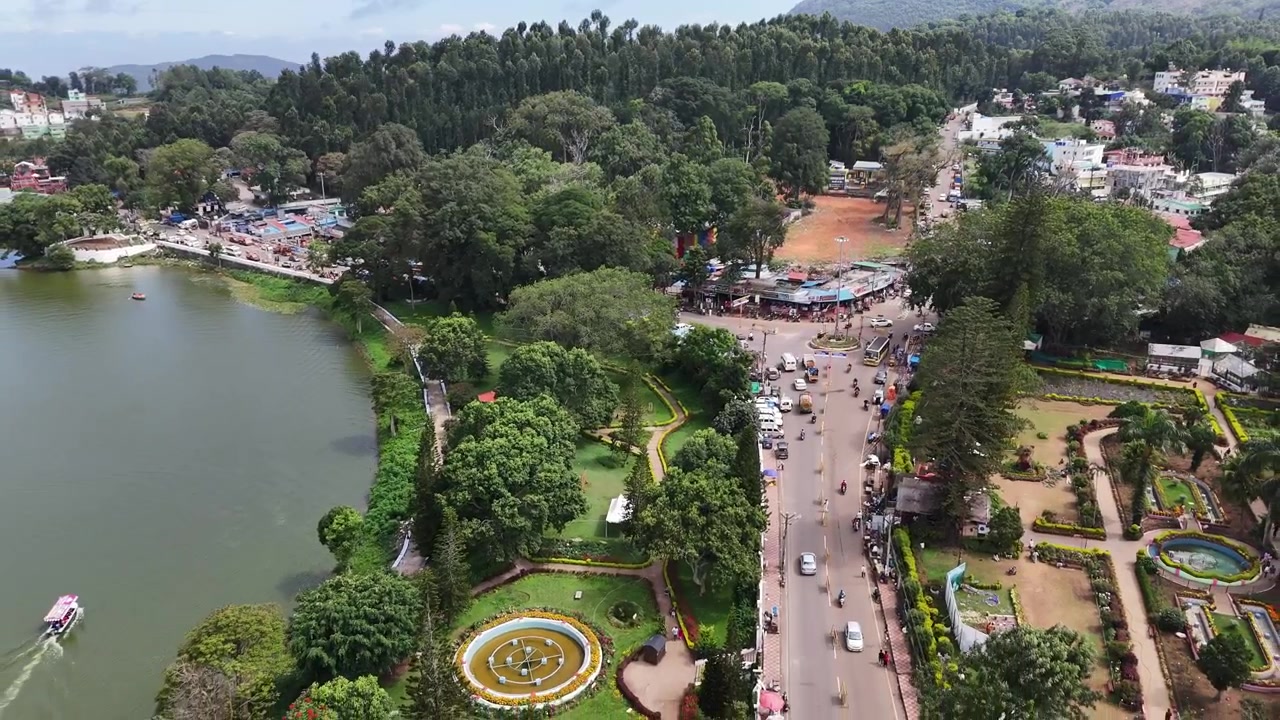 Plynulý zpětný záběr dronu odhalující hlavní křižovatku Yercaud, Smaragdové jezero, upravené zahrady, rušnou dopravu a svěží zeleň. Filmový letecký pohled na horskou stanici v Tamilnádu, Indie